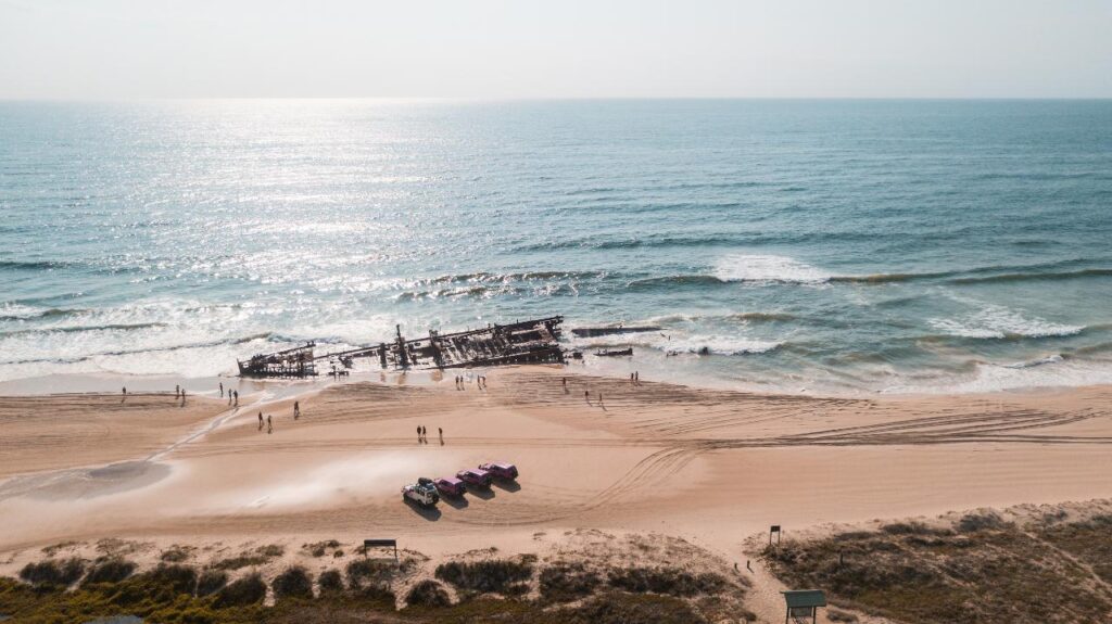 Aerial view of a beach with a shipwreck in shallow water, people walking on the sand, and several vehicles parked near the shoreline.