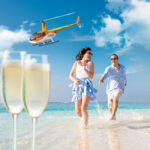 A smiling couple runs along a sandy beach near the ocean, with two glasses of champagne in the foreground and a helicopter flying overhead in a blue sky.
