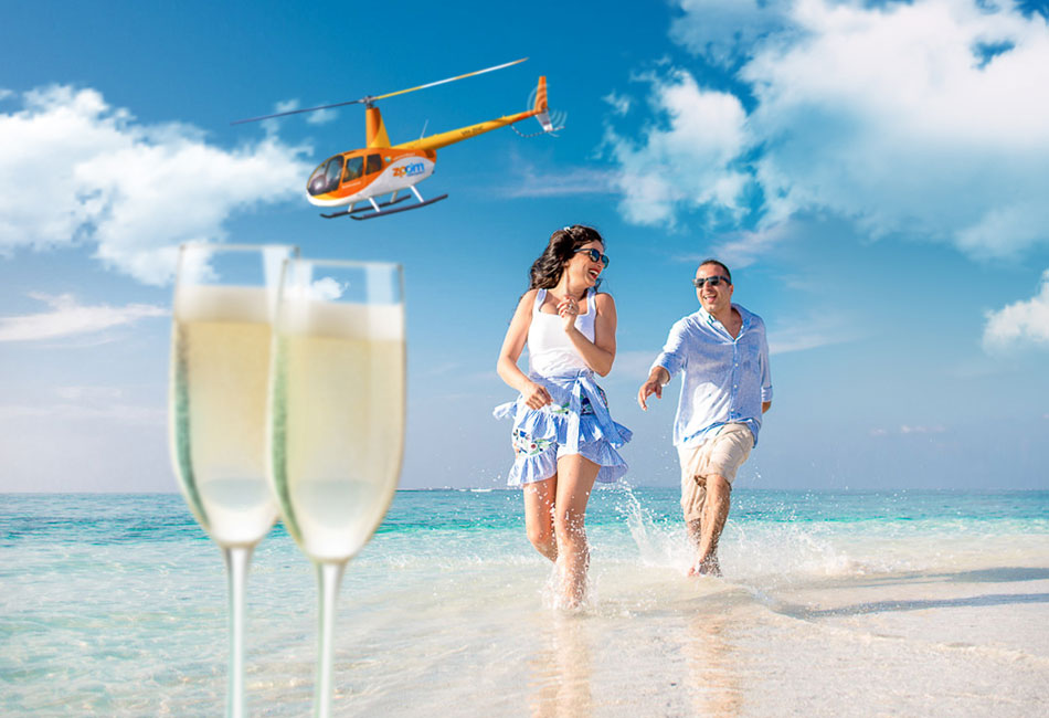 A smiling couple runs along a sandy beach near the ocean, with two glasses of champagne in the foreground and a helicopter flying overhead in a blue sky.