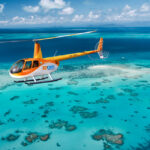 A yellow and orange helicopter flies over clear turquoise water and coral reefs near a small sand island under a partly cloudy sky.
