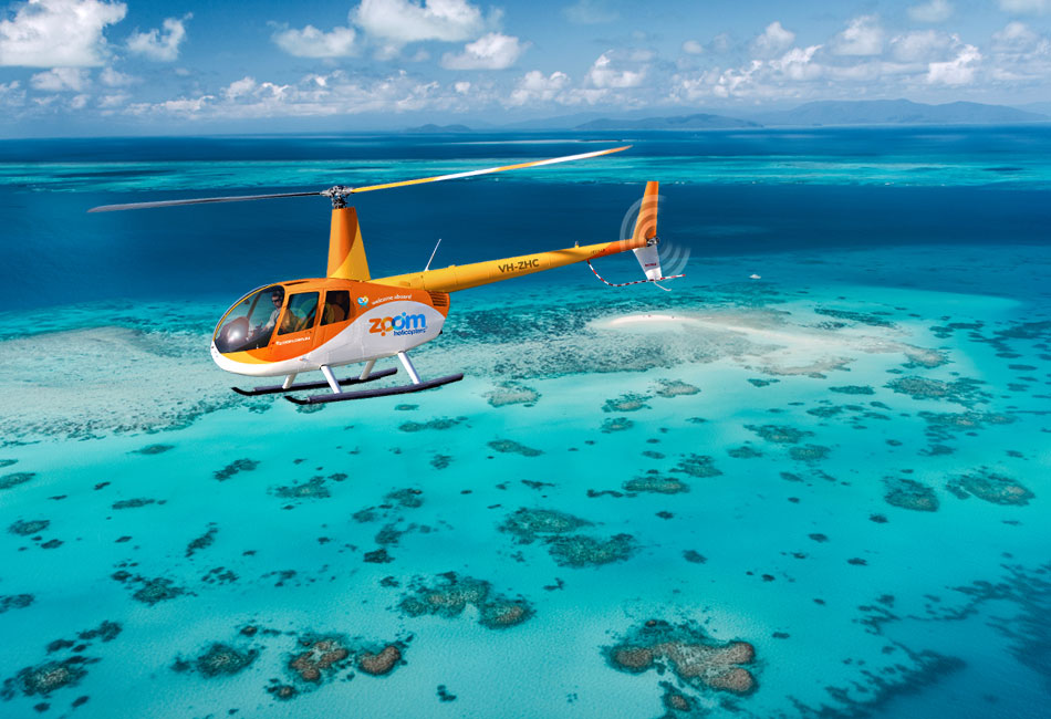 A yellow and orange helicopter flies over clear turquoise water and coral reefs near a small sand island under a partly cloudy sky.