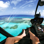 View from inside a helicopter cockpit flying over turquoise ocean waters and coral reefs, with a person's legs and feet visible in the foreground.
