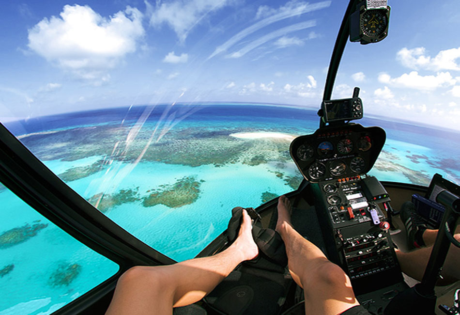 View from inside a helicopter cockpit flying over turquoise ocean waters and coral reefs, with a person's legs and feet visible in the foreground.