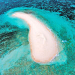 Aerial view of a small, crescent-shaped sandbar surrounded by shallow turquoise water and coral reefs.