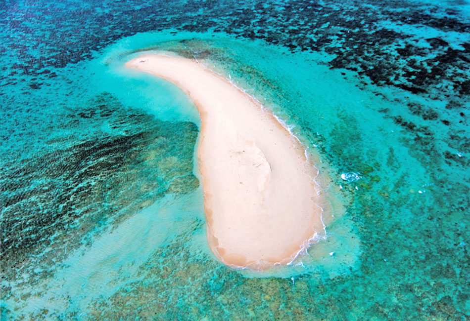Aerial view of a small, crescent-shaped sandbar surrounded by shallow turquoise water and coral reefs.