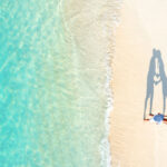 Aerial view of two people lying on a sandy beach near clear turquoise water, casting a shadow that forms a heart shape.