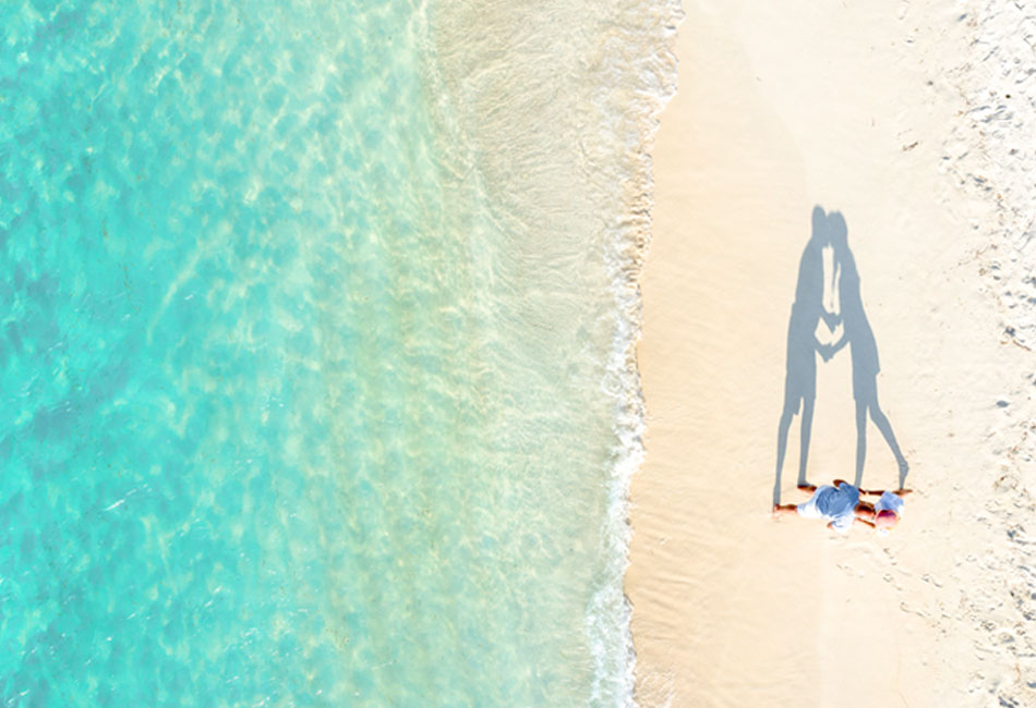 Aerial view of two people lying on a sandy beach near clear turquoise water, casting a shadow that forms a heart shape.