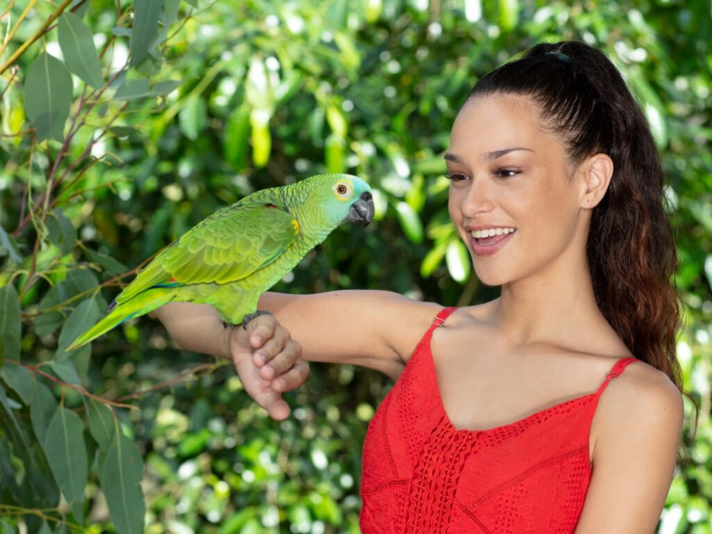 A woman in a red dress smiles while holding a green parrot on her arm, with leafy green foliage in the background.