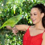 A woman in a red dress smiles while holding a green parrot on her arm, with leafy green foliage in the background.