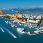 An orange and white helicopter flies over a marina with yachts and boats, with a city and mountains visible in the background under a clear sky.