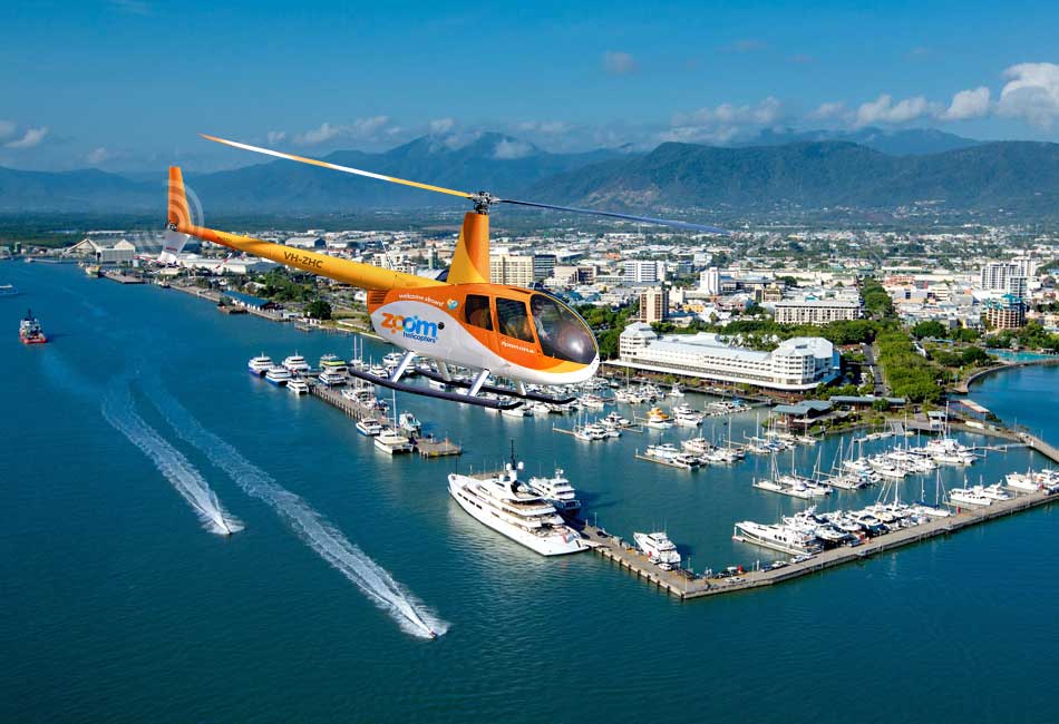 An orange and white helicopter flies over a marina with yachts and boats, with a city and mountains visible in the background under a clear sky.