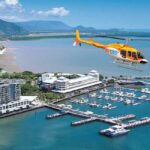 A yellow and orange helicopter flies over a coastal city marina with boats docked, buildings along the shore, and mountains in the background.