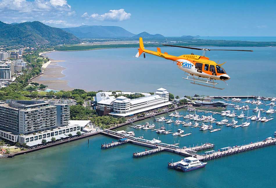 A yellow and orange helicopter flies over a coastal city marina with boats docked, buildings along the shore, and mountains in the background.
