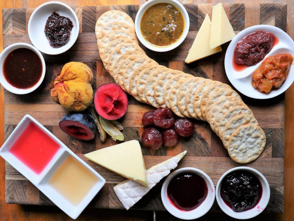 A wooden board with assorted crackers, cheeses, sliced sausage, fresh figs, plums, and small bowls of jams, chutneys, and sauces arranged neatly.
