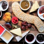 A wooden board with assorted crackers, cheeses, sliced sausage, fresh figs, plums, and small bowls of jams, chutneys, and sauces arranged neatly.