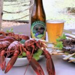 A plate of cooked crawfish, a bottle and glass of beer, and a plate of grilled meat skewers are arranged on a table outdoors near a body of water.