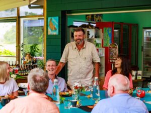 A man stands and speaks while others sit and smile around a table set for a meal in a colorful indoor space.