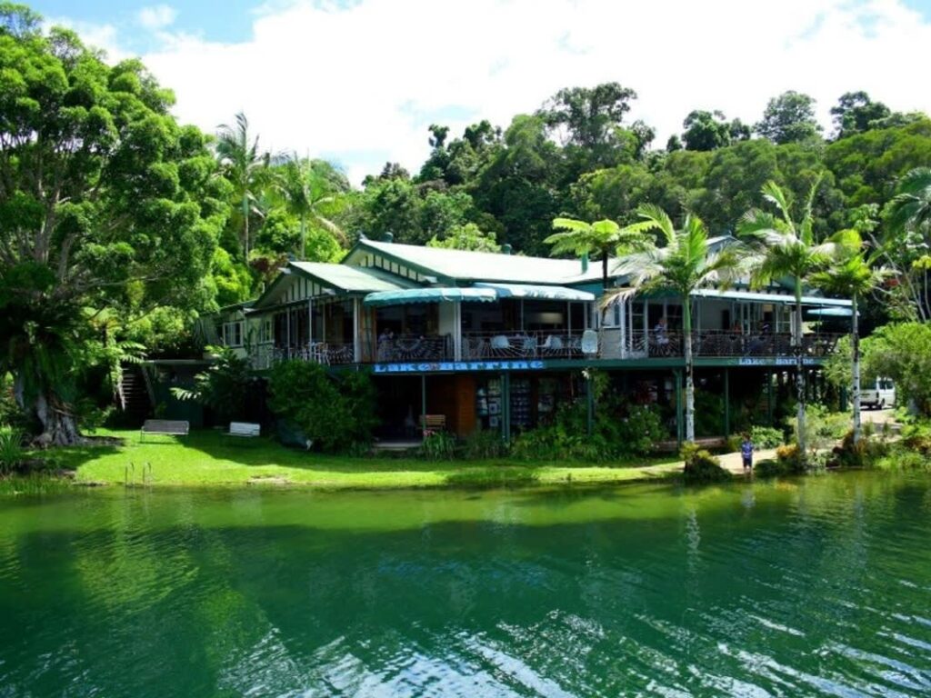 A building with green roofs and open balconies sits next to a calm lake, surrounded by lush trees and greenery under a partly cloudy sky.