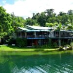 A building with green roofs and open balconies sits next to a calm lake, surrounded by lush trees and greenery under a partly cloudy sky.