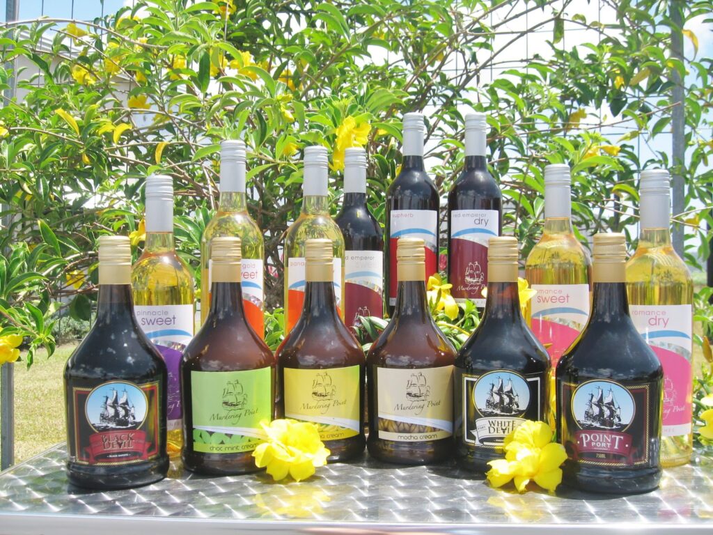 Bottles of assorted wines and liqueurs are arranged on a metal surface outdoors, with yellow flowers and green foliage in the background.