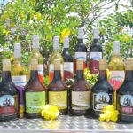Bottles of assorted wines and liqueurs are arranged on a metal surface outdoors, with yellow flowers and green foliage in the background.